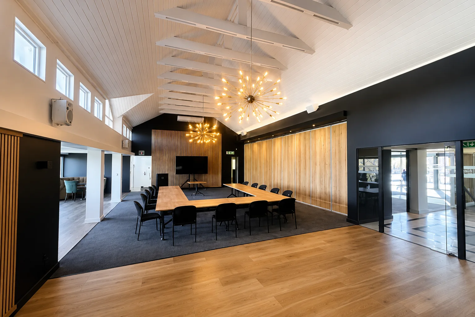 Spacious conference room with U-shaped wooden tables, black chairs, high white ceiling with beams, pendant lights, and wood panelled walls